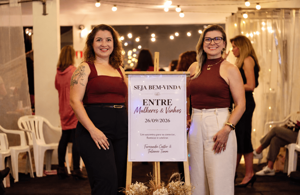 Entre Mulheres & Vinhos promove encontro de autoconhecimento em São Leopoldo Duas participantes posam ao lado de um cartaz do encontro Entre Mulheres & Vinhos