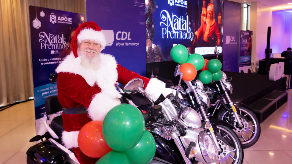 Natal Premiado 2026 da CDL-NH terá sorteio de motos e iPhones Papai Noel sobre uma motocicleta, com balões verdes e laranjas e painéis da campanha Natal Premiado da CDL Novo Hamburgo ao fundo.