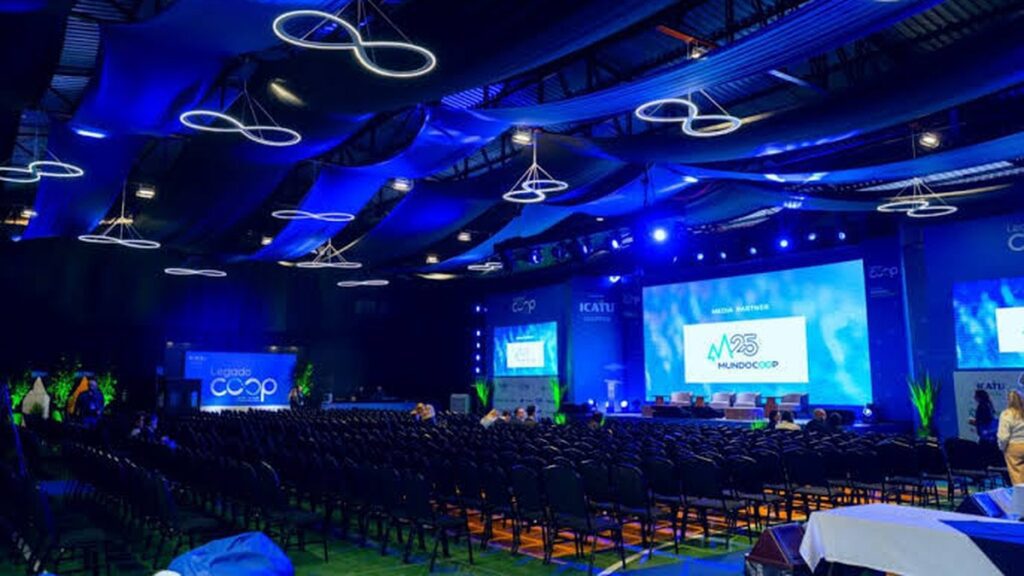 Legado Coop 2027 terá espaço para cooperativas e programação de imersão cultural Auditório do Legado Coop com palco, telões e iluminação azul
