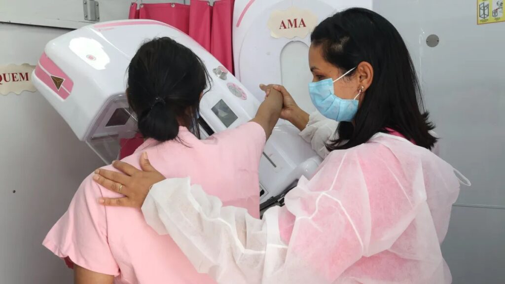 Profissional de saúde auxilia mulher posicionada em aparelho de mamografia para detecção do câncer de mama