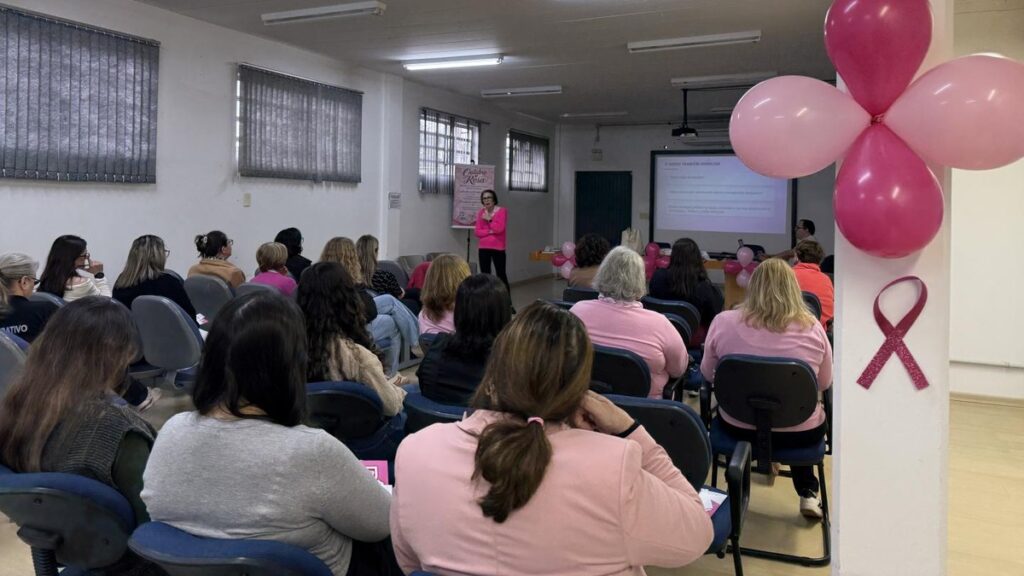 Público acompanha palestra do Outubro Rosa em Estância Velha, em sala decorada com balões e laço rosa.