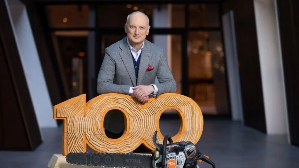 Centenário da STIHL é tema de encontro empresarial em São Leopoldo Representante da STIHL posa atrás de uma escultura de madeira com o número 100